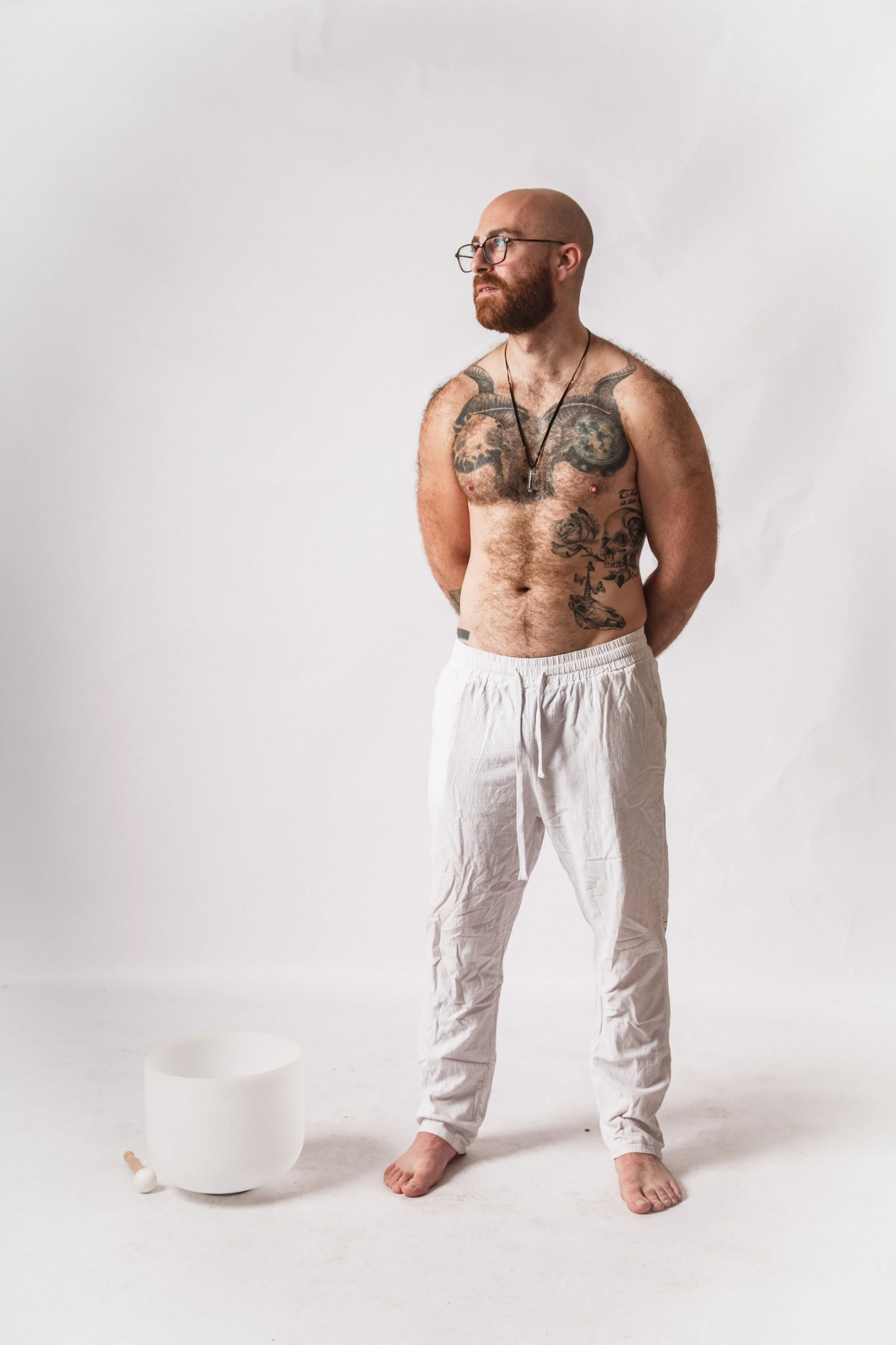 A man with a beard, wearing glasses, white pants, and no shirt, stands against a plain white background. He has tattoos on his chest and abdomen, and wears a necklace. His hands are behind his back, and he is barefoot. There is a white bowl, a mallet, and a small object on the ground in front of him.