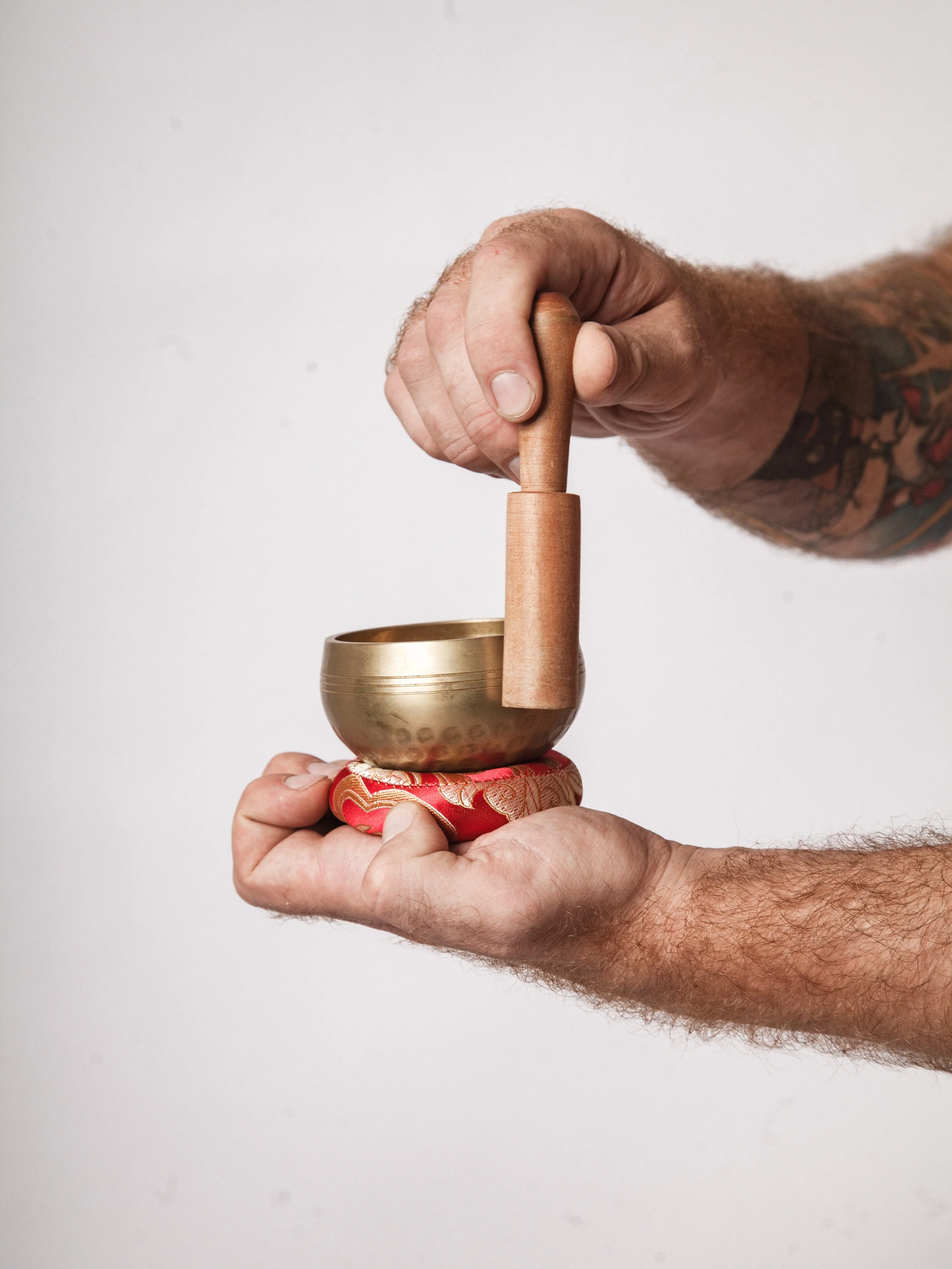 A person's hand holding a singing bowl with a wooden mallet, with a red decorative cloth underneath, against a plain white background.