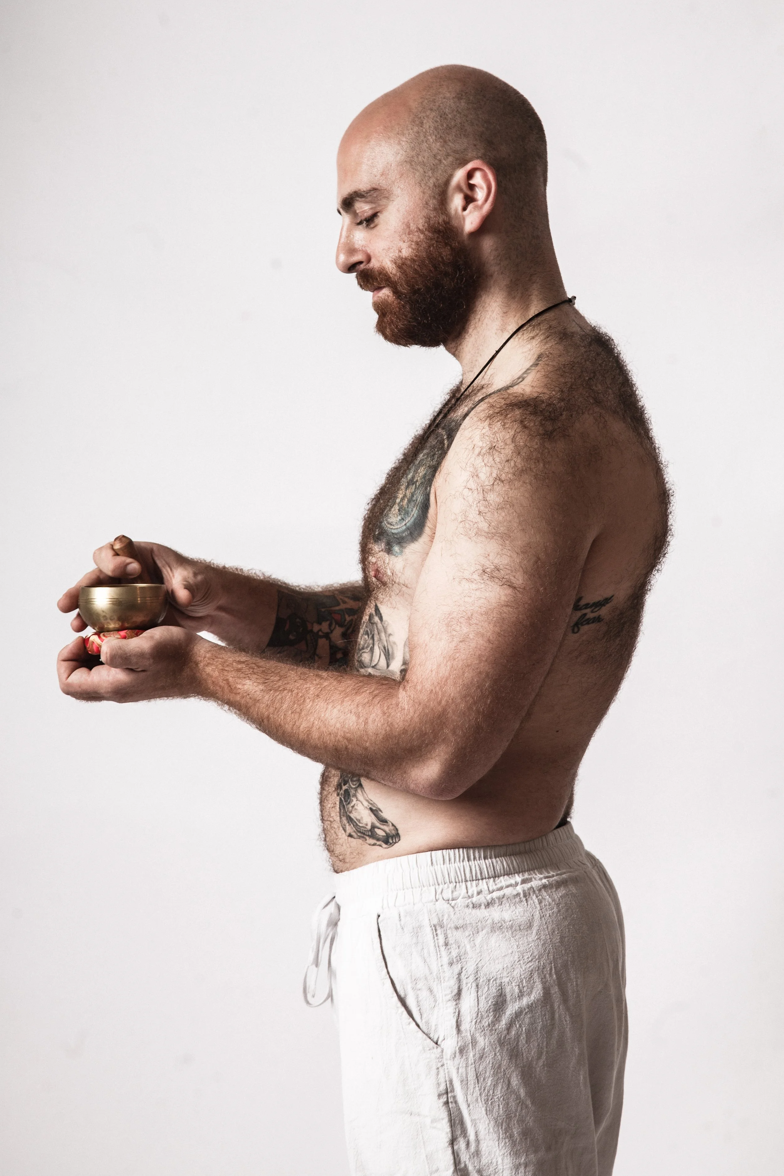 A shirtless man with a beard and tattoos on his chest and arm, wearing light-colored sweatpants, stands in profile against a plain white background, holding a small bowl.