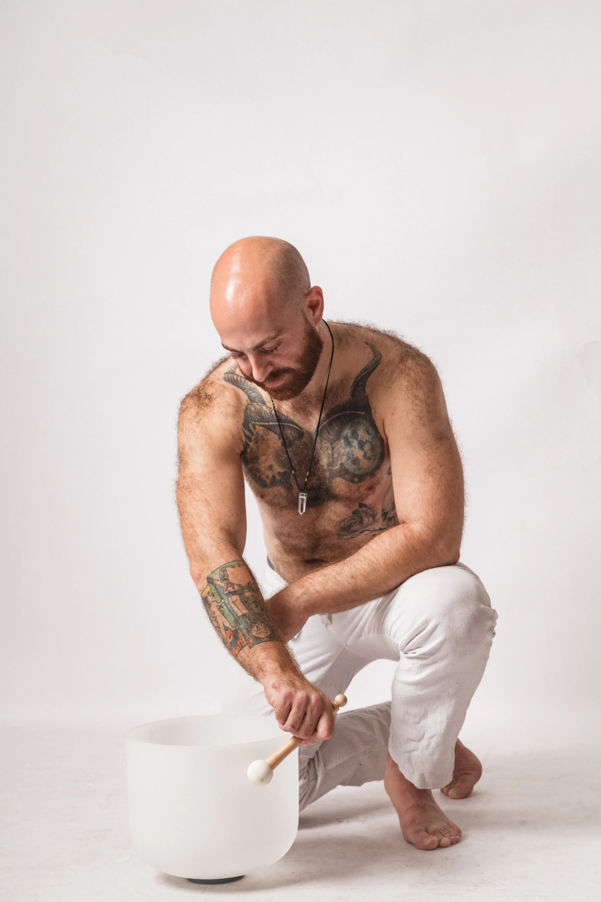 A tattooed man with a bald head and beard is crouched down and holding a mallet, about to strike a tuning fork placed on a white round object, against a plain white background.