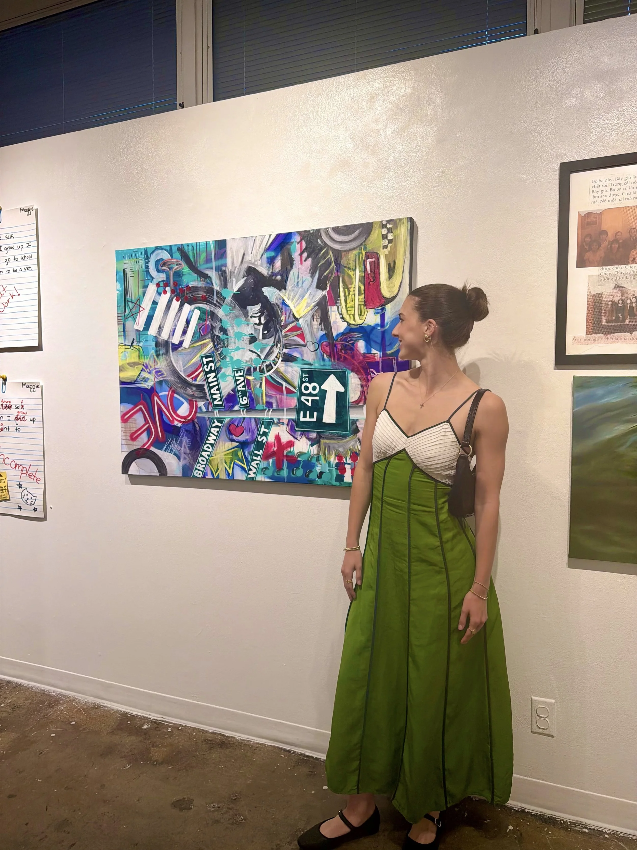 A woman wearing a green dress with a striped top design and black shoes, standing in front of colorful street art on a white gallery wall.