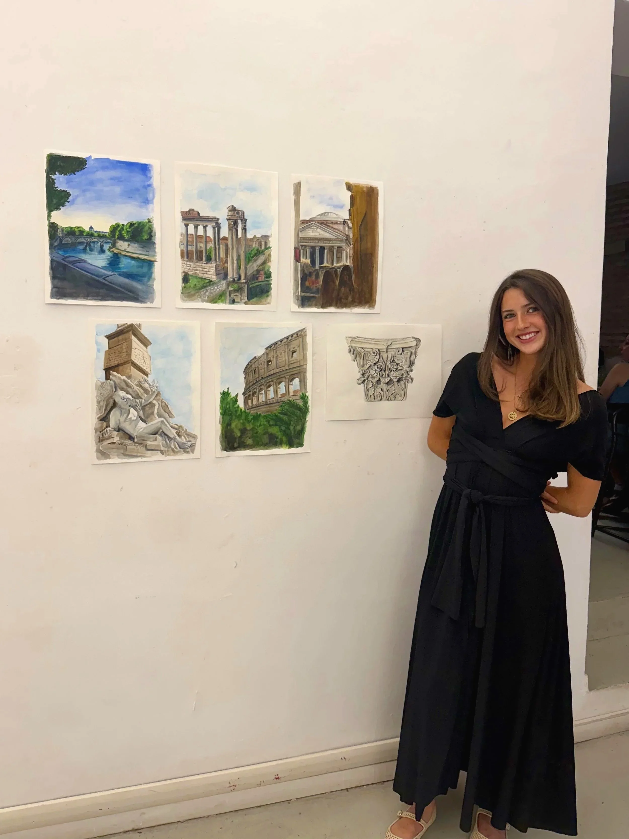 A woman in a black dress with a pleated skirt and strappy shoes is standing next to a white wall with watercolor paintings of Roman ruins and architecture. She is smiling and has long brown hair.