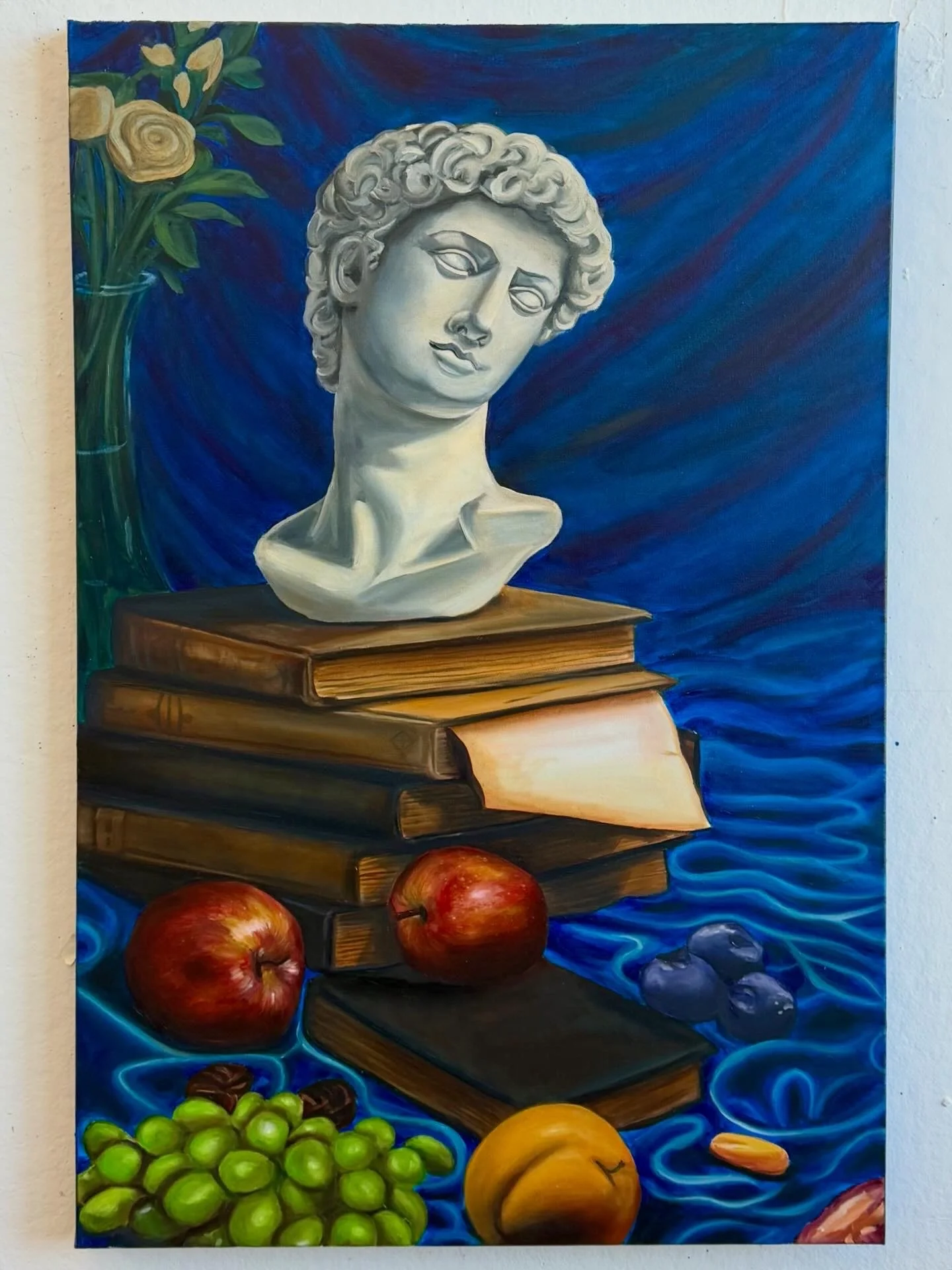 A still life painting of a classical marble bust on a stack of antique books with fruits including apples, grapes, and a peach, set against a blue background with a vase of yellow roses.