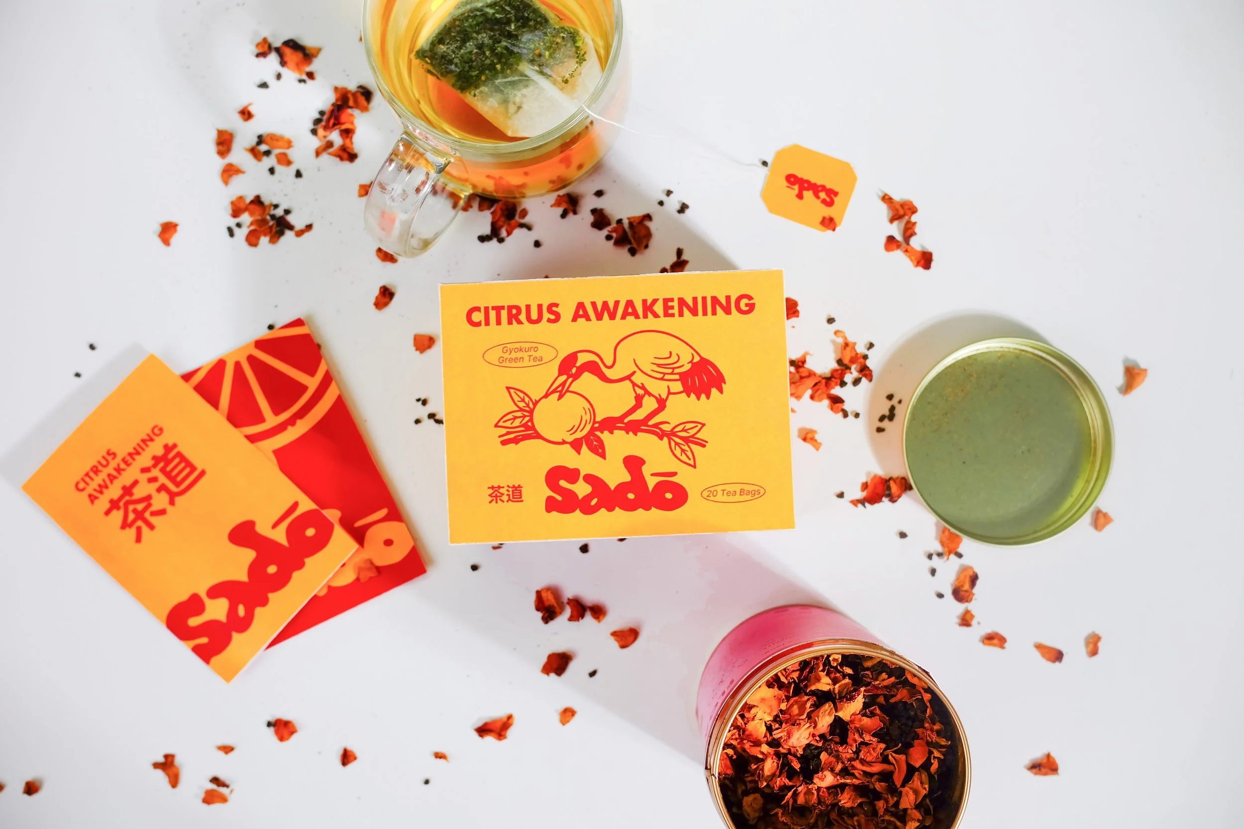 A top-down view of a tea setup with orange and yellow safflower tea packaging labeled 'Citrus Awakening' and 'Sadō', loose safflower petals, and two cups of tea, one with green tea and the other with a darker variant, on a white surface.