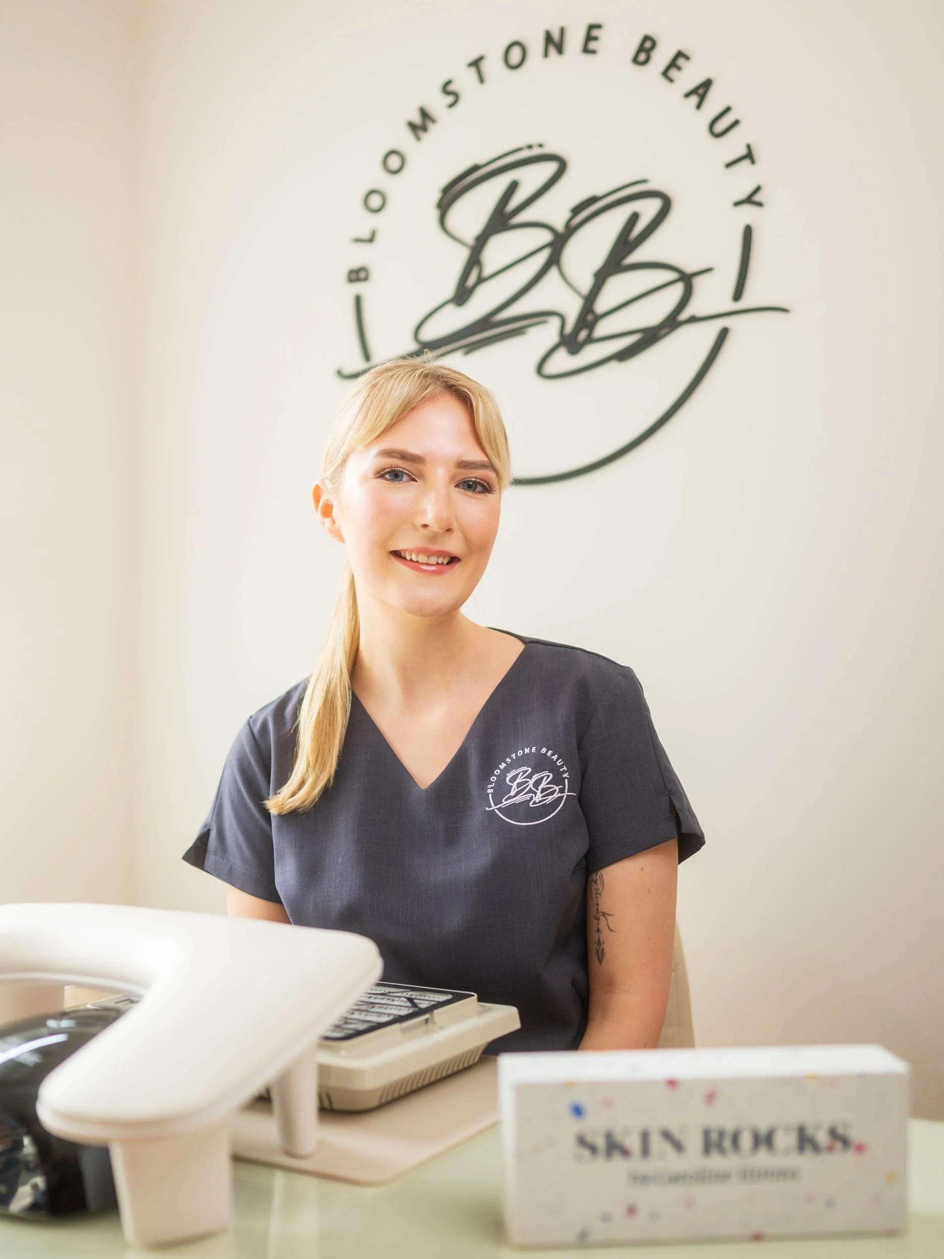A woman with blonde hair and a tattoo on her arm smiling, sitting at a table with a book titled 'Skin Rocks' and a calculator, in front of a wall with a logo that reads 'Bloomstone Beauty'.