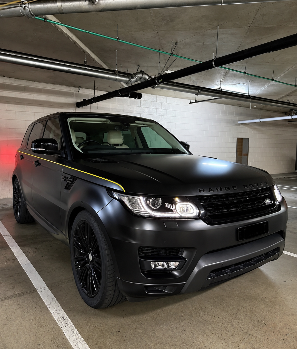 A black Range Rover SUV parked in an underground parking garage with a matte finish, black wheels, and a yellow stripe along the side.