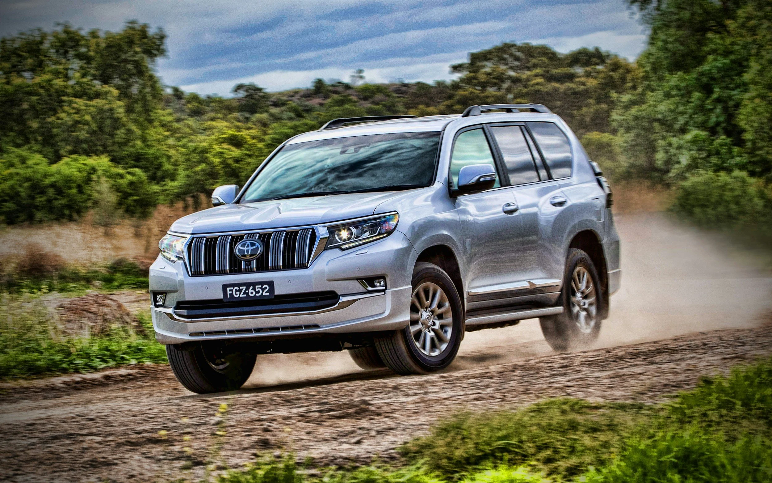 A silver Toyota SUV driving on a dirt road surrounded by trees and green foliage under a cloudy sky.