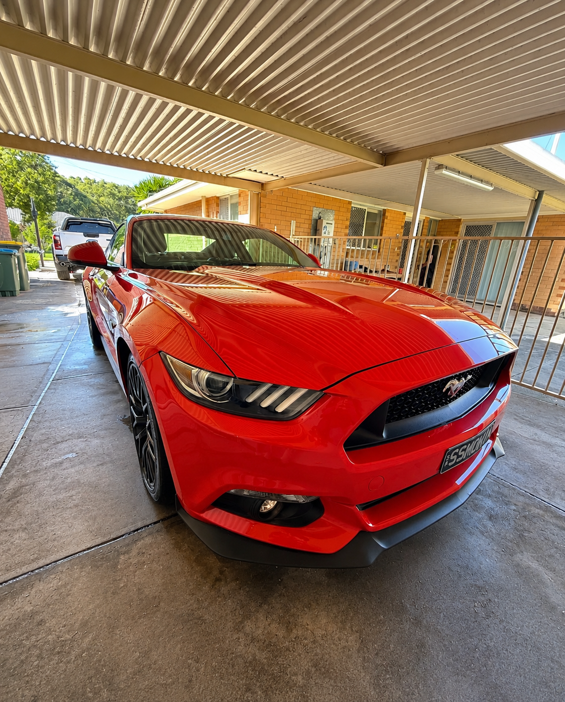 Red Ford Mustang car parked in a covered driveway of a residential building with brick walls and metal railings.