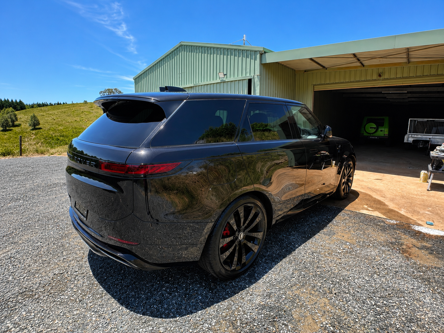 Black Range Rover parked outside a green building with an open garage, on a gravel driveway with a grassy hill in the background and a clear blue sky.