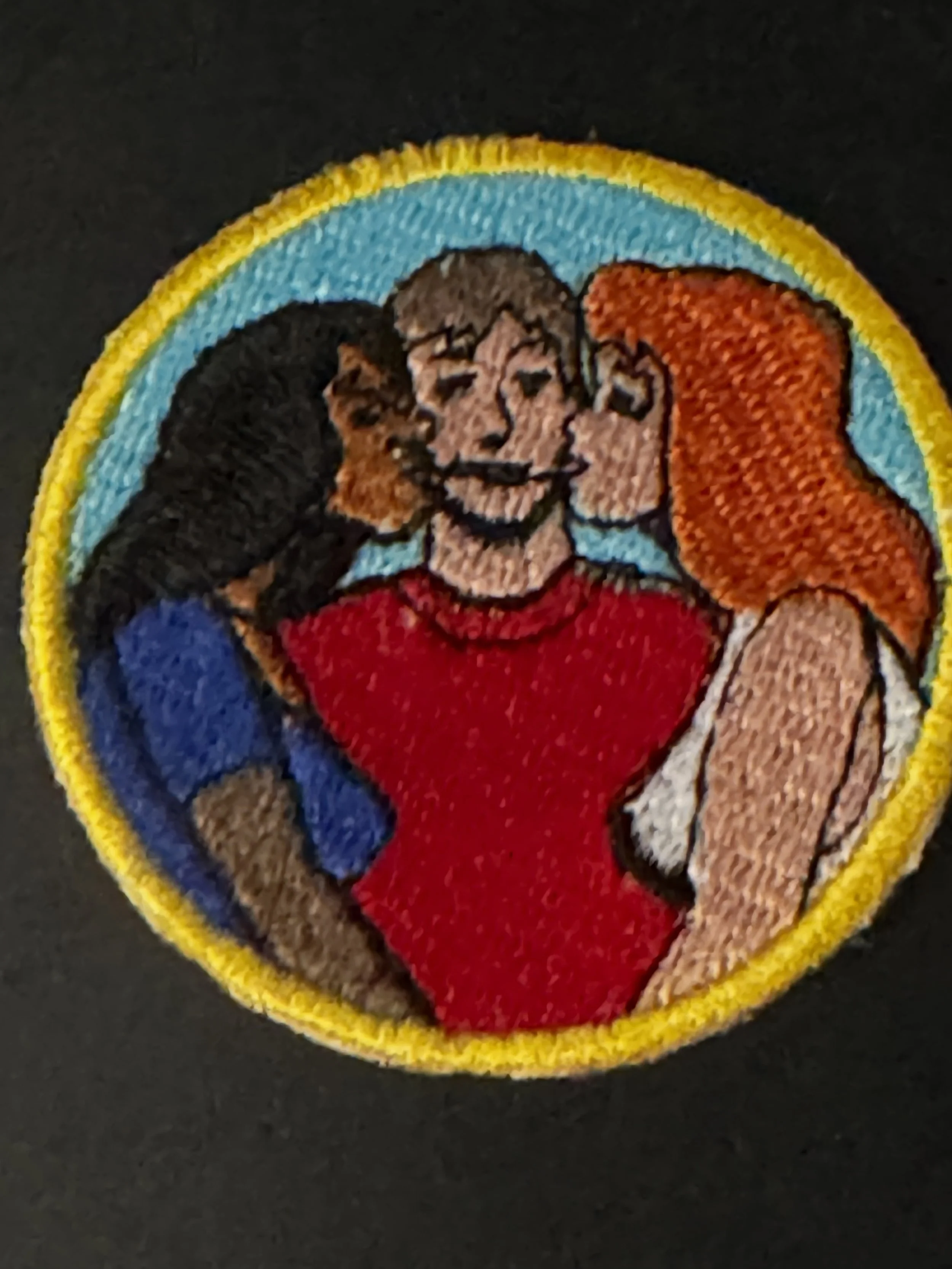 An embroidered patch featuring four women with different hair styles and clothing, framed with a yellow border and a light blue background.