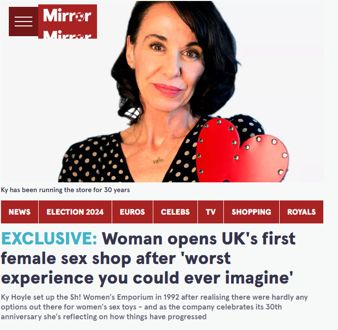 Woman with shoulder-length dark hair holding a red heart-shaped object, wearing a black top with white polka dots, in front of a newspaper website featuring headlines about a woman opening the UK's first female sex shop.