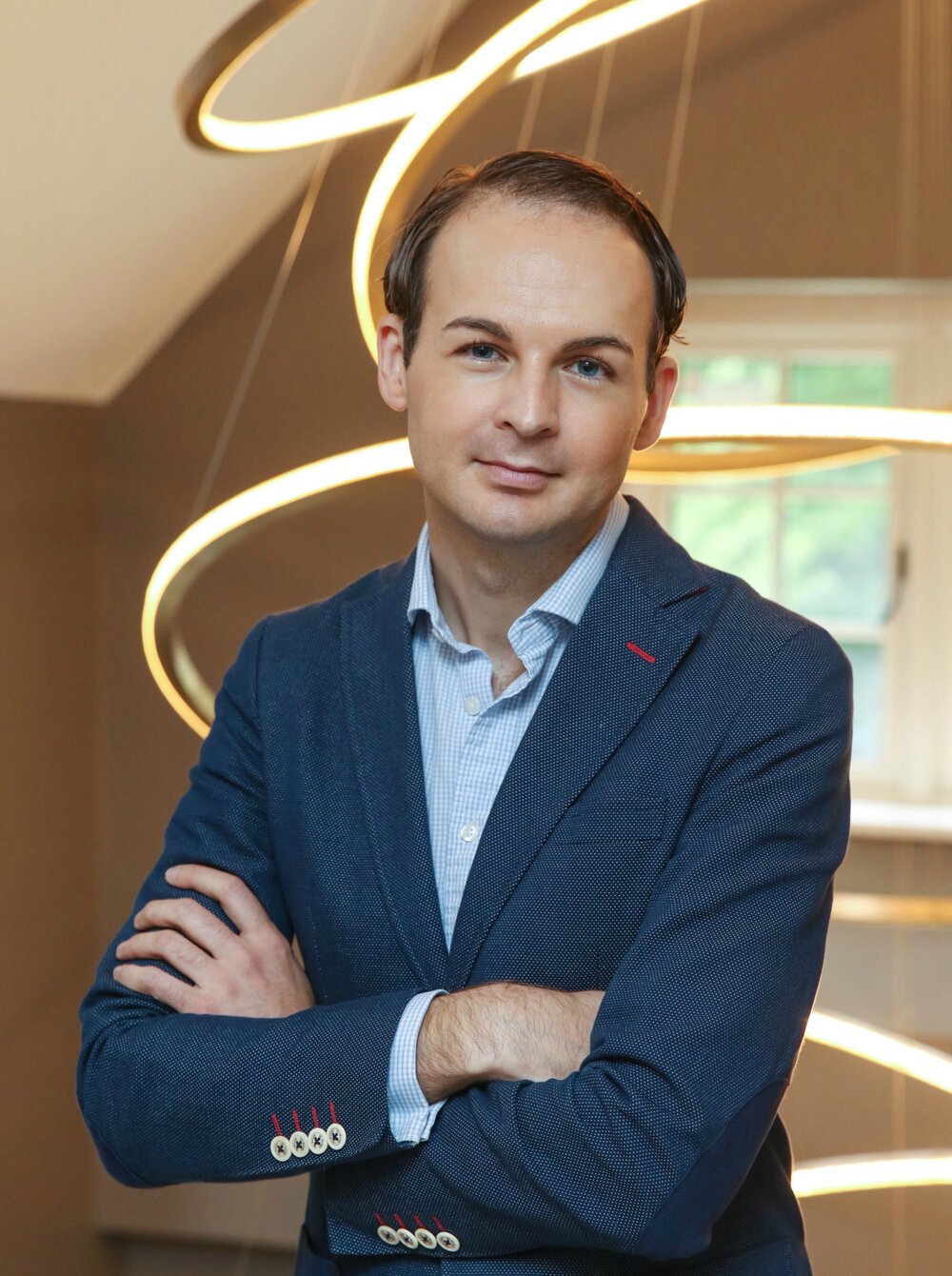 A man in a blue blazer and dress shirt with crossed arms, standing indoors with modern lighting and a window in the background.