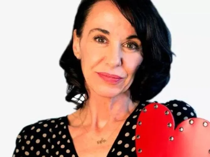 A woman with dark hair wearing a black polka dot top holding two red hearts with metal studs, smiling at the camera.