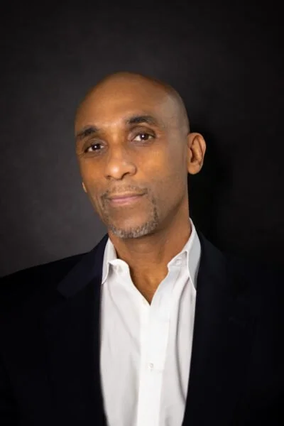 A man with a shaved head and goatee, wearing a white dress shirt and dark blazer, standing against a dark background.