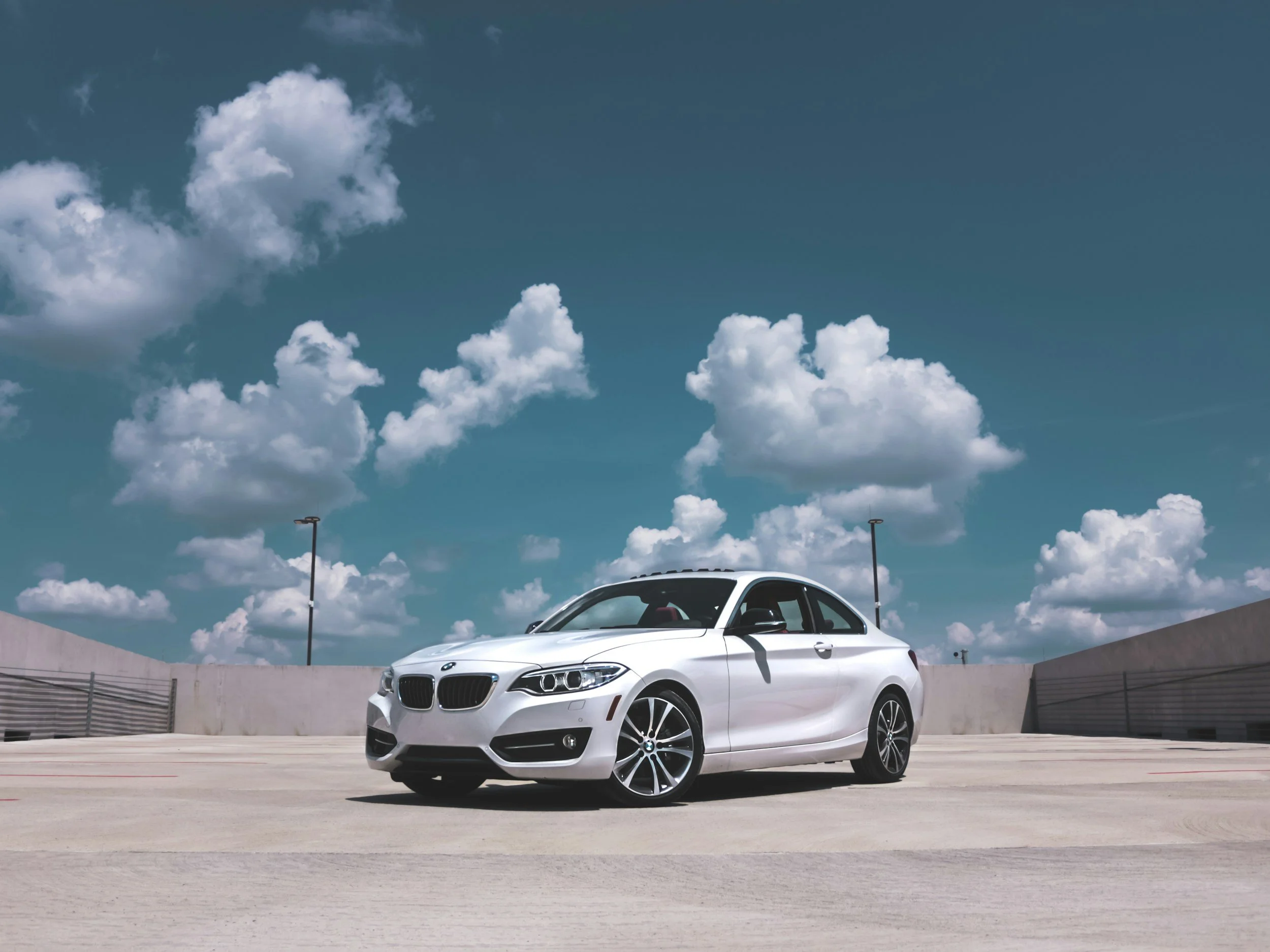 A white BMW car parked in an outdoor parking lot under a blue sky with scattered clouds. The parking lot has a concrete wall and a metal fence, with some street lamps visible in the background.