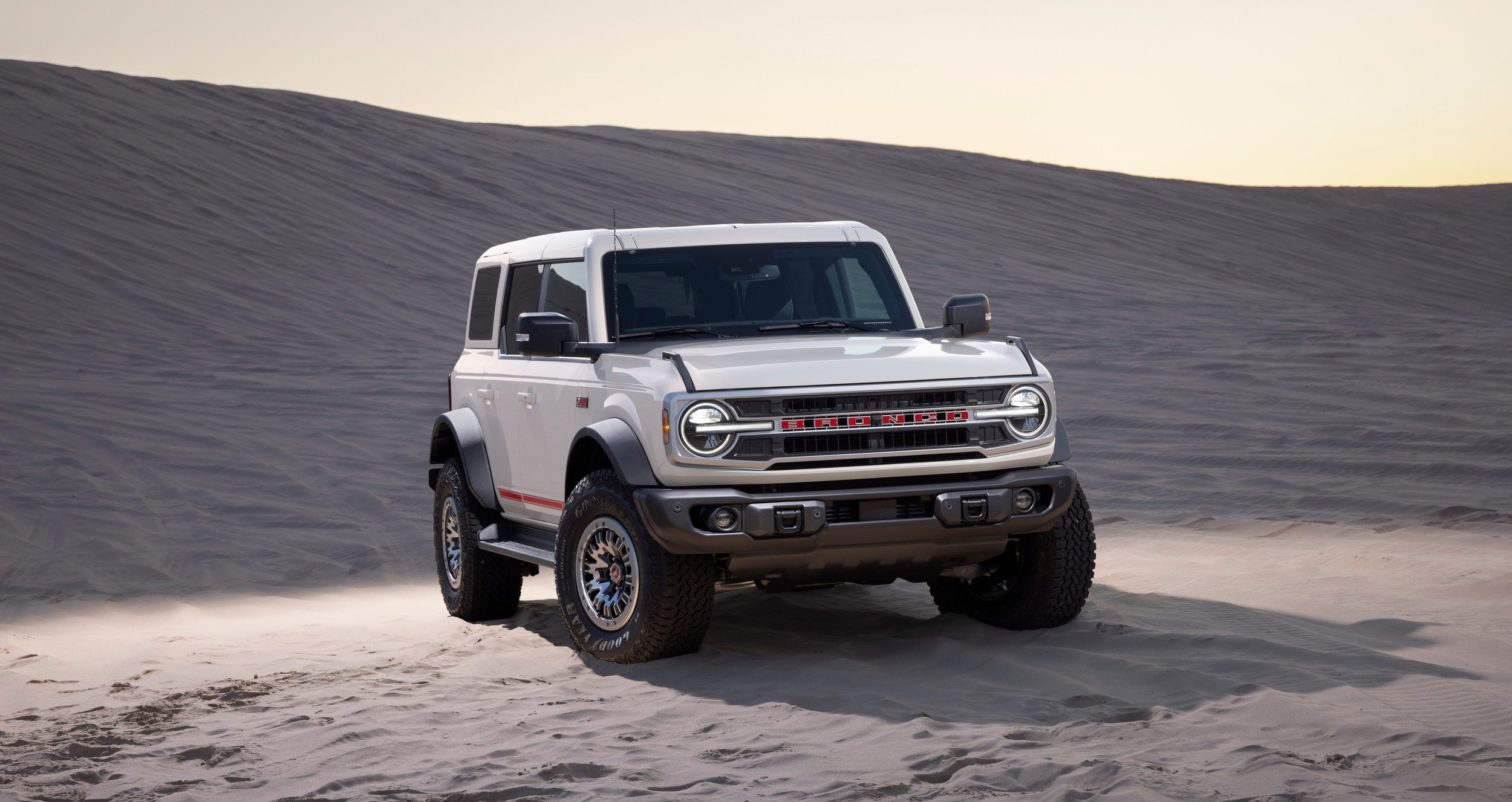 A white off-road vehicle driving through sandy desert terrain at sunset with rolling sand dunes in the background.