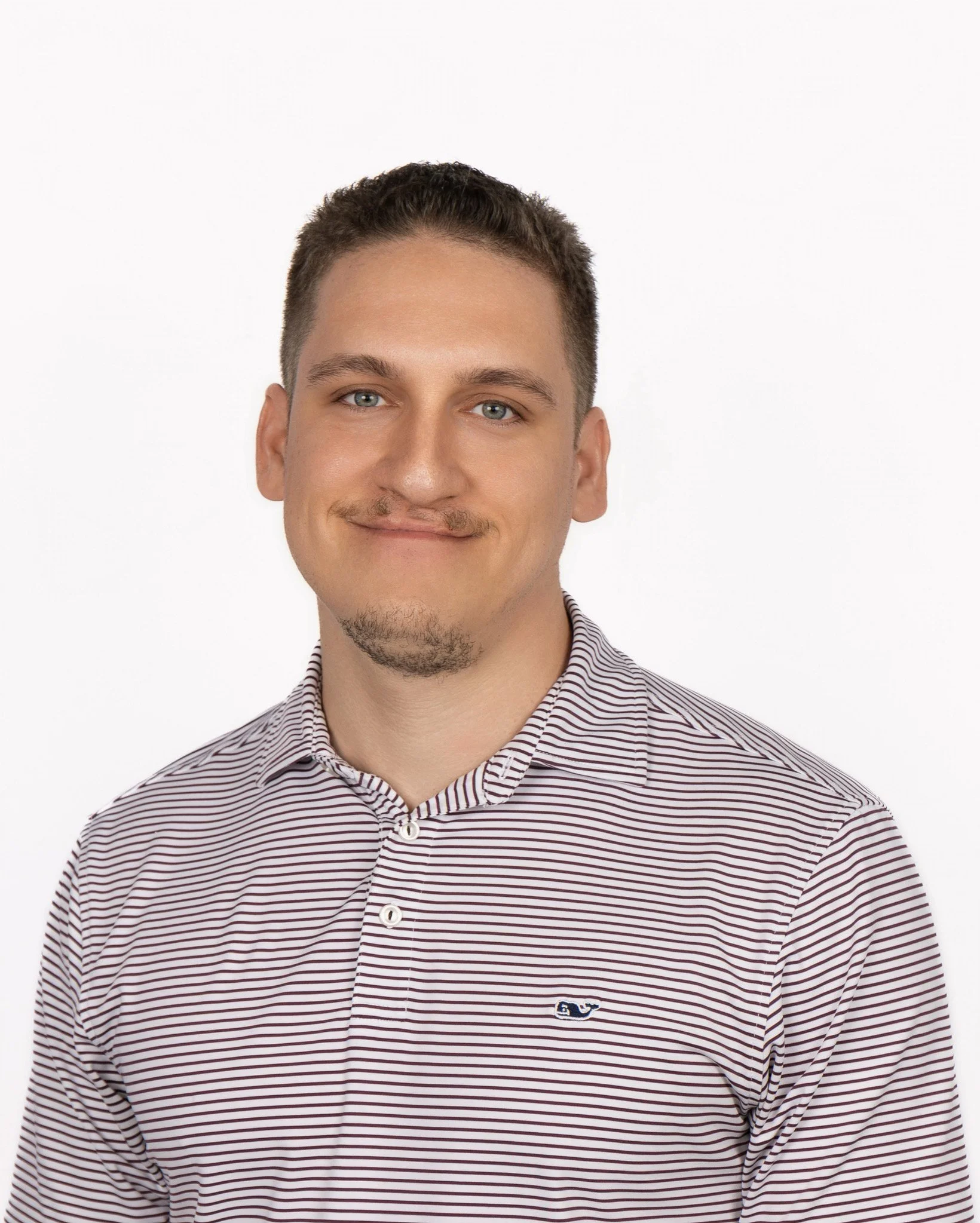 A young man with short brown hair, light blue eyes, and a slight smile, wearing a striped button-up shirt, standing against a plain white background.