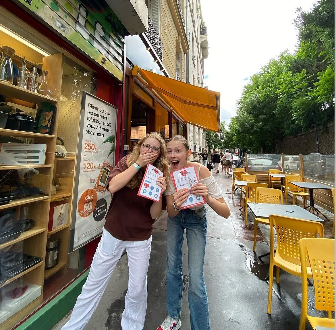 Deux jeunes femmes souriantes et surpris, tenant des menus, debout devant un café avec des tables jaunes à l'extérieur, avec des arbres et des voitures en arrière-plan, un jour pluvieux.
