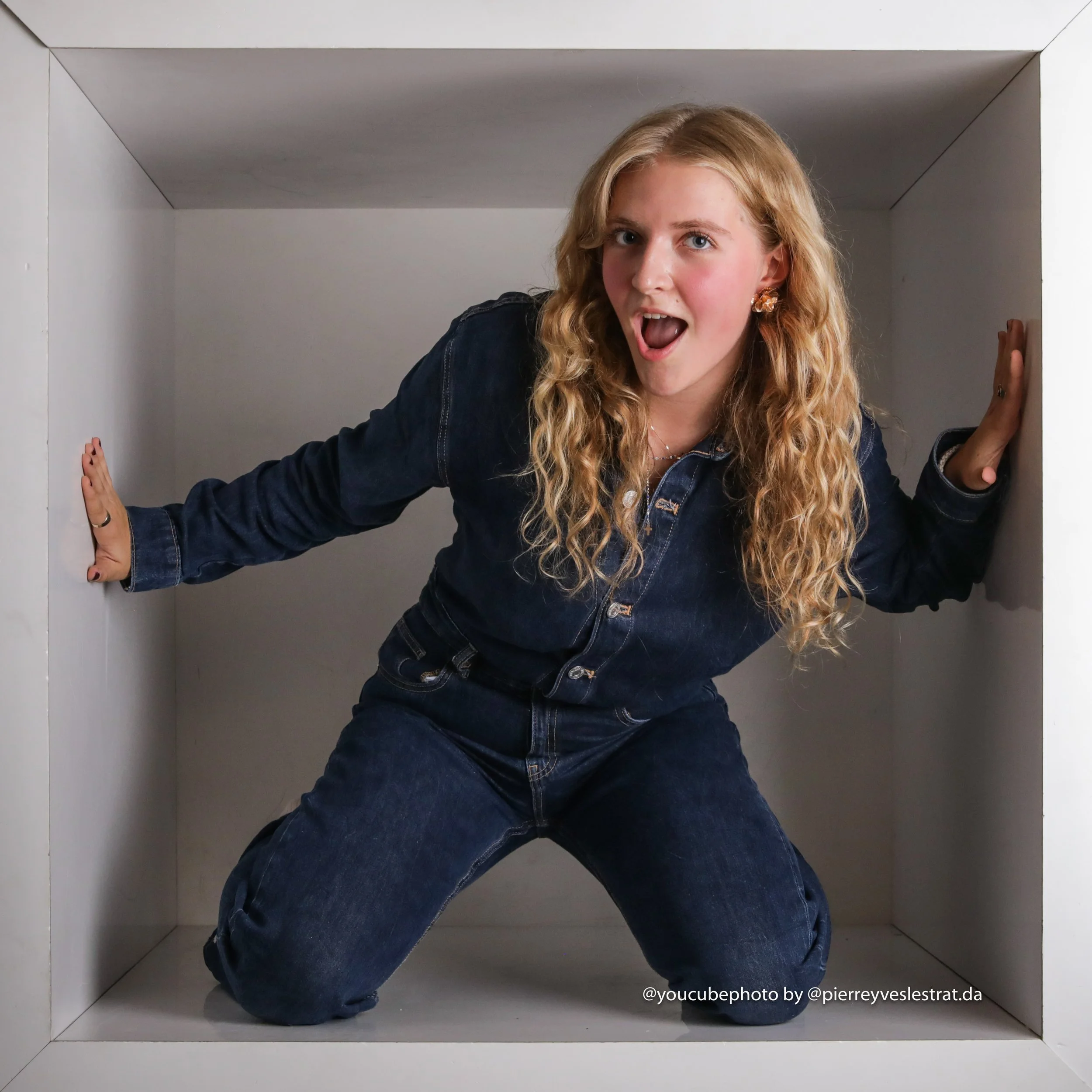 Jeune femme aux cheveux blonds et longs, portant un jean, posant dans une petite cabine photo blanche, en montrant un visage surpris et une expression joyeuse.