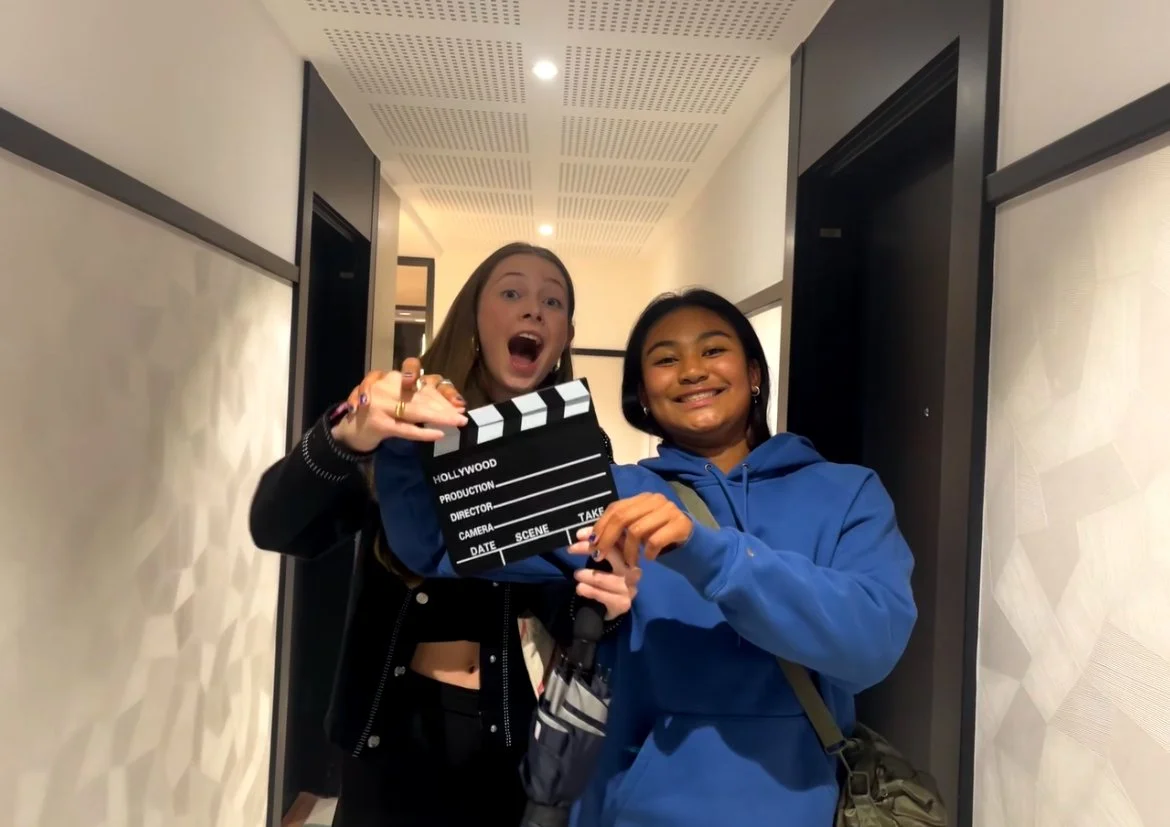 Deux jeunes femmes souriantes, tenant une clap de cinéma, dans un couloir intérieur.