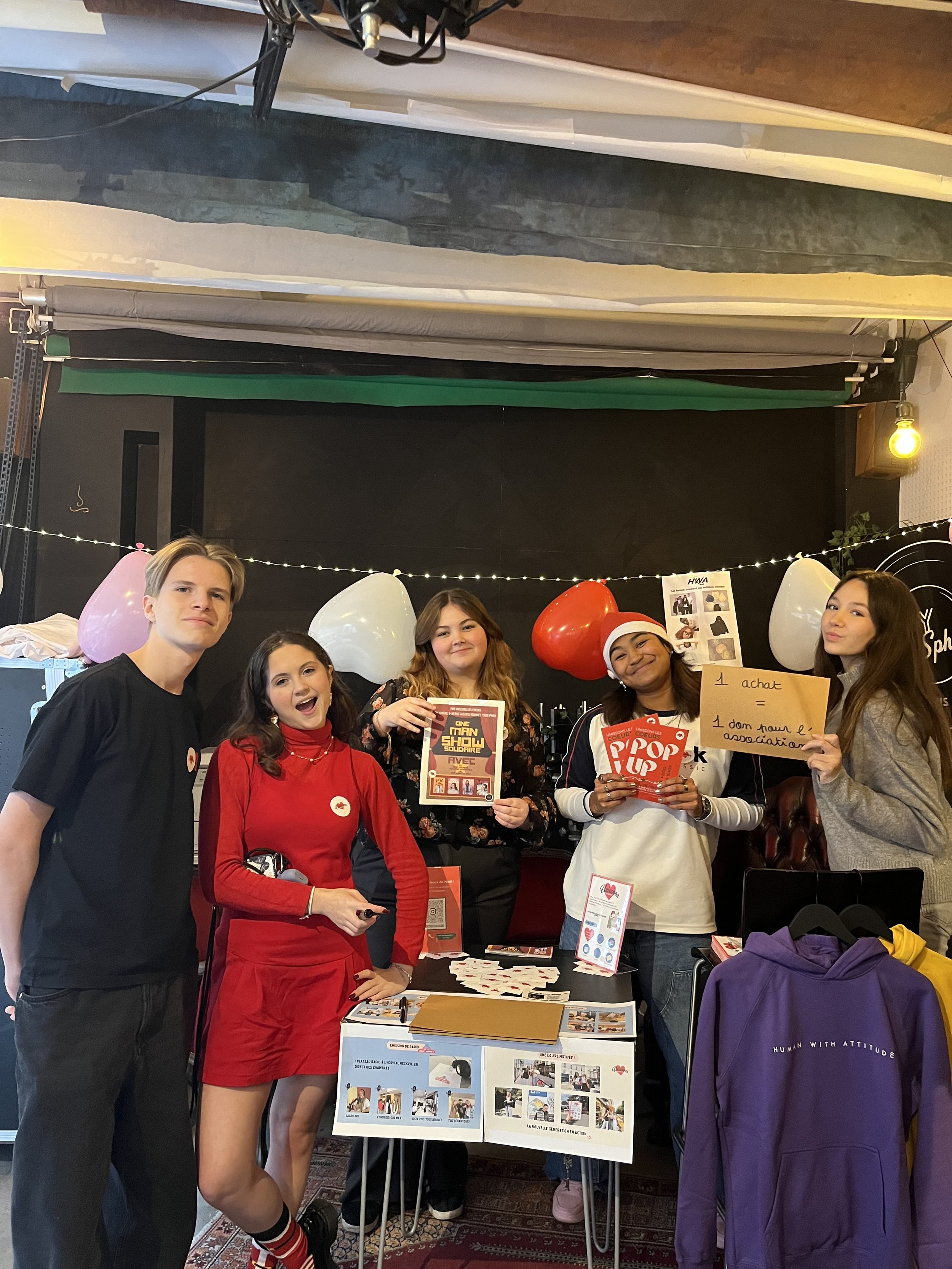 Groupe de jeunes gens posant et présentant une affiche lors d'un événement caritatif ou d'une campagne de sensibilisation, avec des Ballons et un stand, tous souriants.