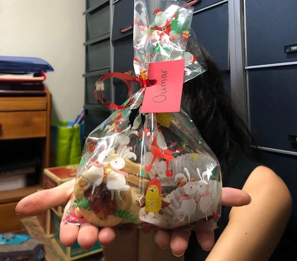 Un cadeau de Noël emballé dans un sac en plastique transparent avec des décorations de Noël, porté par une personne. Le paquet contient des figurines de lapins et d'oiseaux en papier ou en bois.