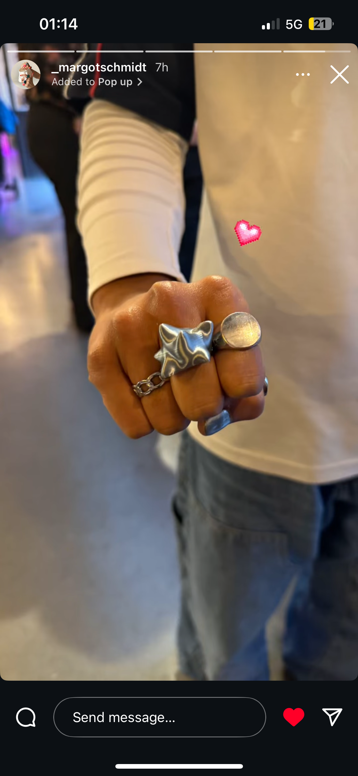 Une main avec plusieurs bagues, dont une en forme de nuage, portée au centre de l'image, avec un fond flou de personnes et d'un espace intérieur