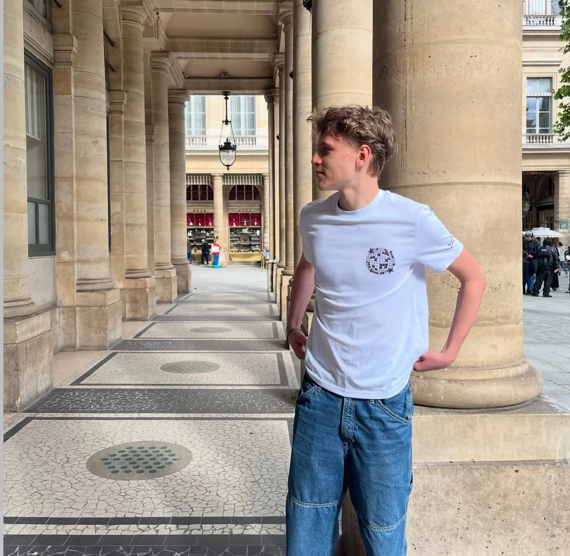 Jeune homme en t-shirt blanc et jeans, debout à côté d'une colonne en pierre, sous une architecture classique à Paris, avec une boutique en arrière-plan et des passants.