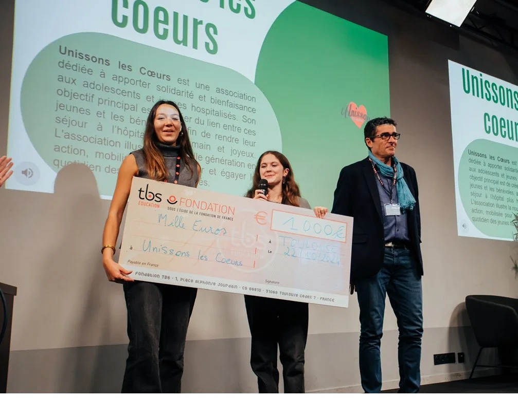 Trois personnes sur scène lors d'une cérémonie de remise de prix, avec un grand chèque de 1000 euros au centre, sous un projecteur, avec un fond vert et un écran affichant un texte en français sur une association nommée 'Unissons les Cœurs'.