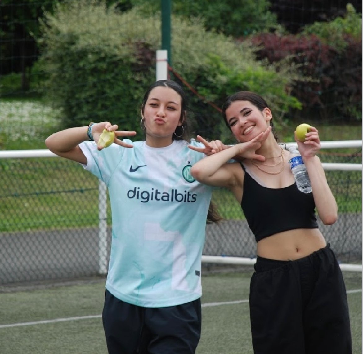 Deux jeunes femmes souriantes posant avec des pommes en main sur un terrain de sport, l'une portant un maillot de football, l'autre une tenue décontractée. En arrière-plan, une clôture et des arbres.