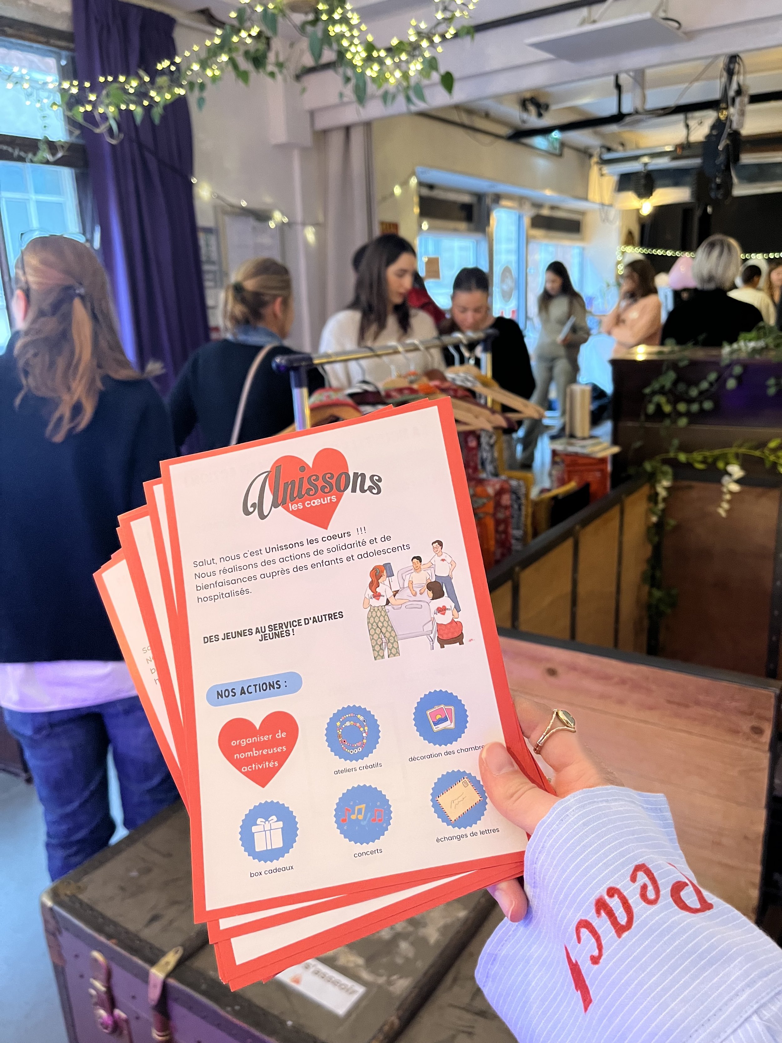 Une main tenant un flyer rouge pour une organisation appelée Unissons les cœurs, avec des détails sur leurs actions caritatives. En arrière-plan, on voit des personnes dans une salle décorée avec des guirlandes lumineuses bleues et vertes, au sein d’un lieu intime ou événementiel.