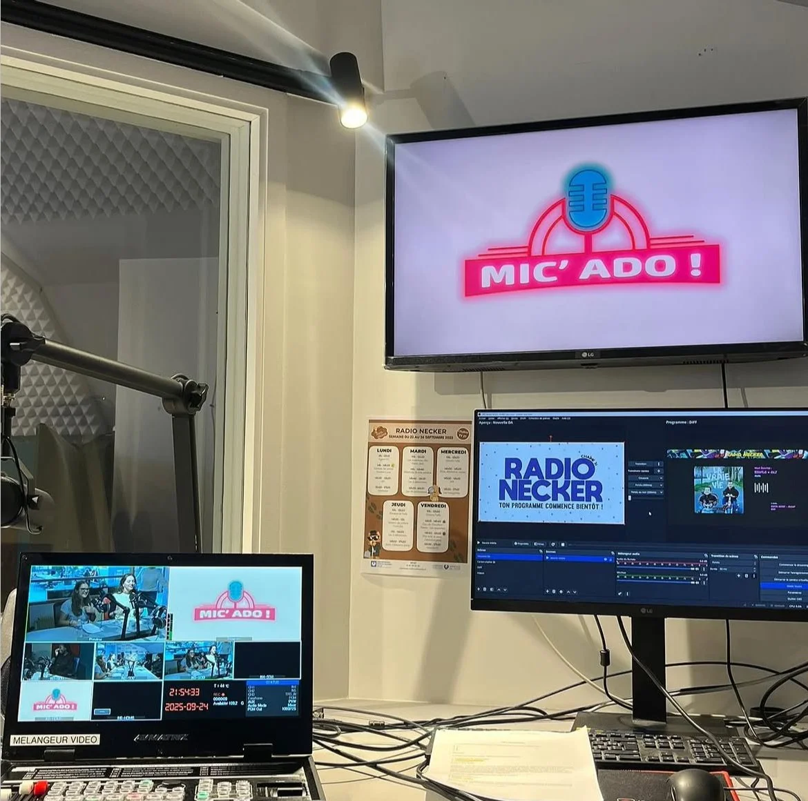 Studio de radio avec plusieurs écrans affichant des logos et un plateau d'enregistrement, une affiche avec le programme de la semaine, et deux femmes en studio lors d'une émission.
