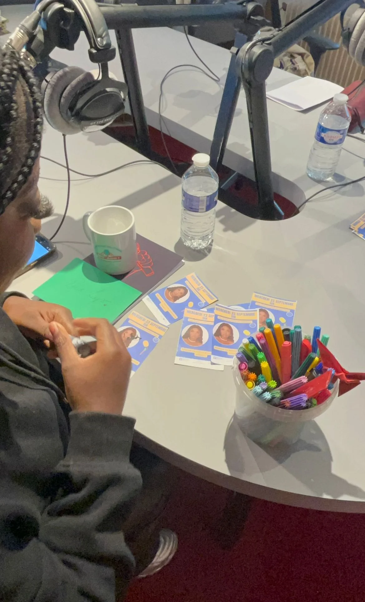 Une personne assise à une table avec des flyers, un mug, une bouteille d'eau, des crayons de couleur et un casque audio; un bureau avec un micro et un ordinateur en arrière-plan.