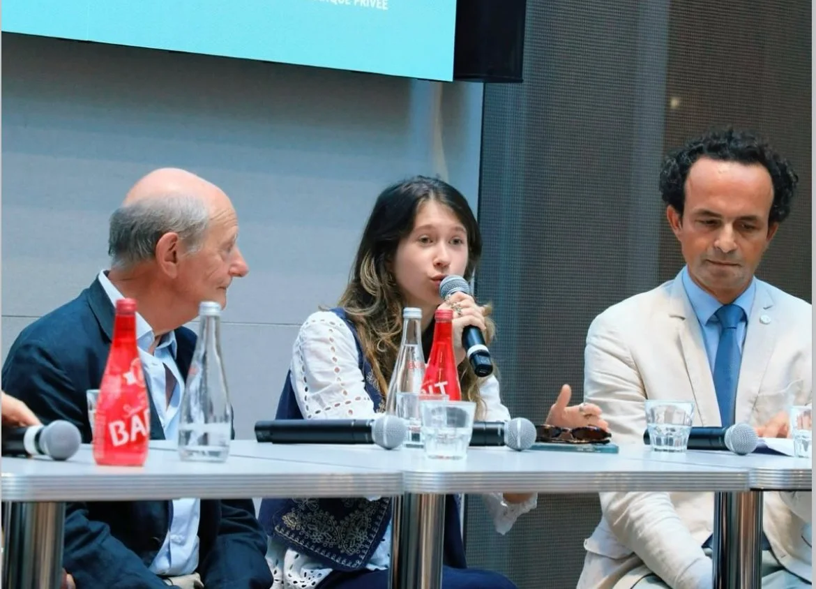 Trois personnes assises à une table lors d'une conférence ou d'un panel, dont une jeune femme parlant dans un micro, avec des bouteilles d'eau et des microphones devant elles.