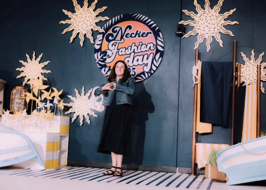 Femme posant devant une décoration de style plage à l'occasion d'un événement de mode à Necker Fashion Day, avec des étoiles et des soleils en décorations, et un panneau indiquant l'événement.
