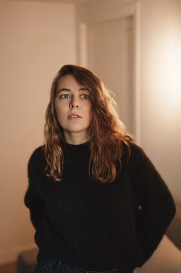 Jeune femme aux cheveux bouclés et roux, portant un pull noir, dans une pièce intérieure avec une lumière douce