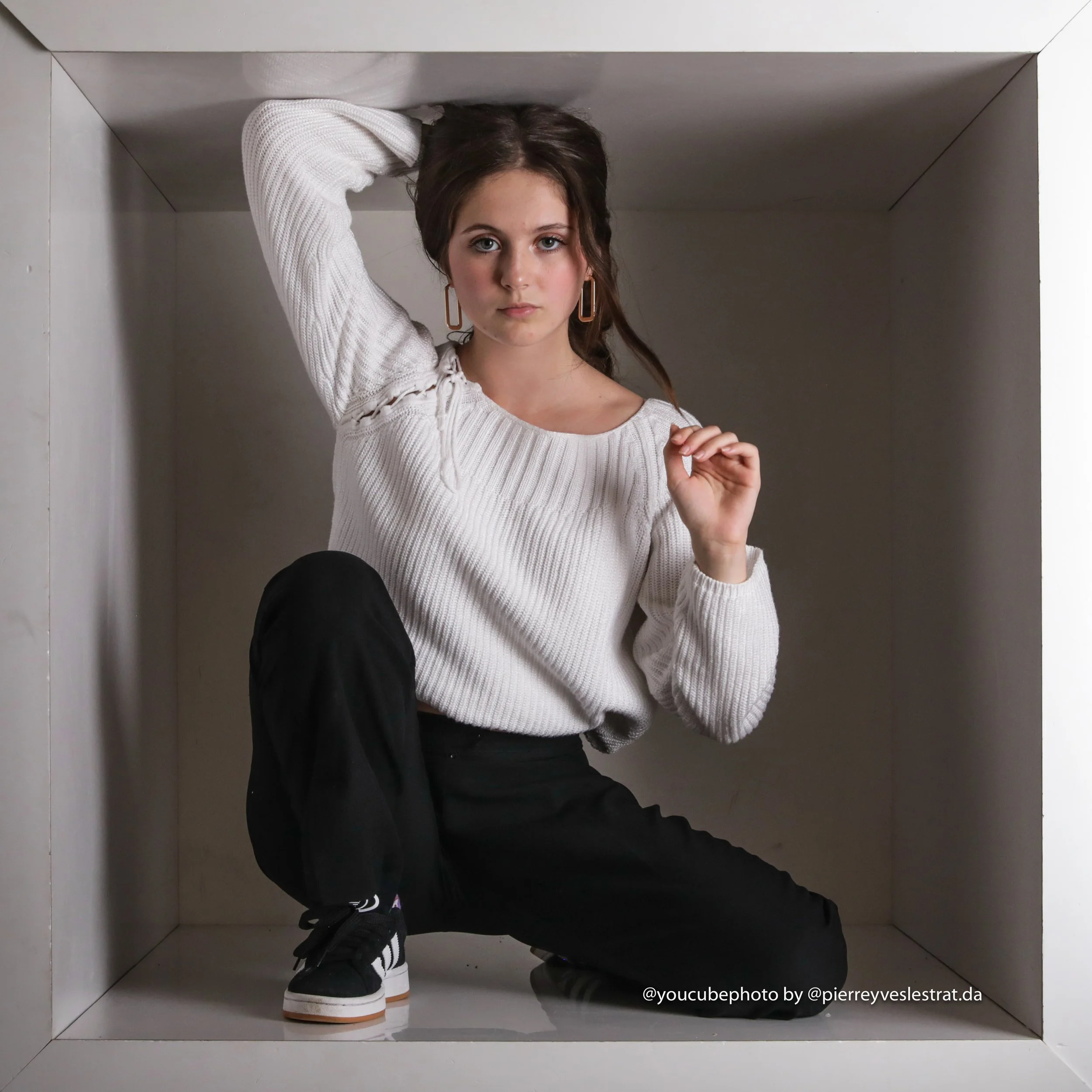 Jeune femme à l'intérieur d'une cabine d'essayage, portant un pull blanc et des baskets noires, avec des boucles d'oreilles longues, tenant sa tête de la main gauche et tombée sur le côté gauche.