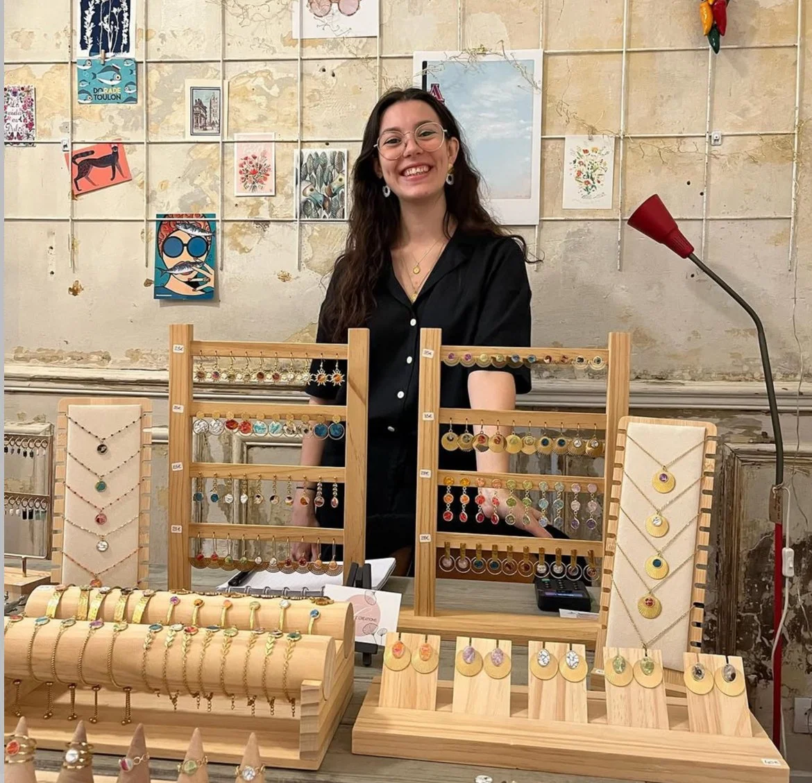 Femme souriante derrière un stand de bijoux colorés exposés sur des présentoirs en bois, avec un mur décoratif en arrière-plan. Elle porte des lunettes et des boucles d'oreilles, en intérieur avec une ambiance artistique.