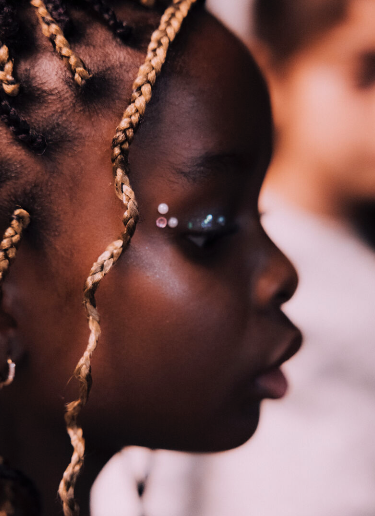 Portrait d'une jeune femme noire avec de petites tresses brunes, maquillage avec des paillettes autour de l'œil, regardant vers la droite.