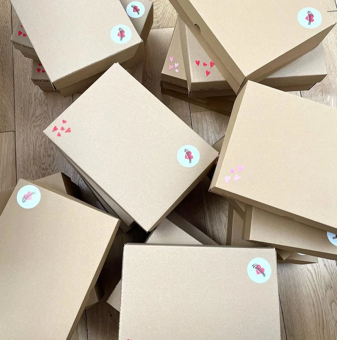 Several cardboard boxes with pink heart stickers and round stickers with a pink beverage logo on a wooden floor.