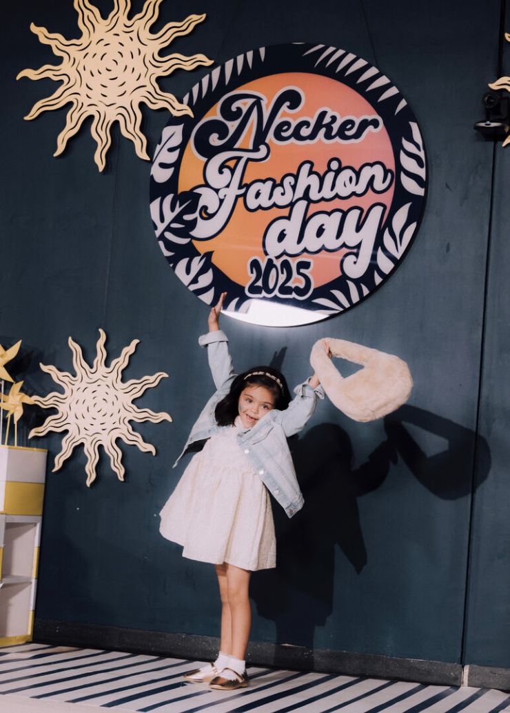Jeune fille souriante posant devant un mur avec un logo de la journée de la mode Necker 2025, décorations en soleil en bois autour du logo.