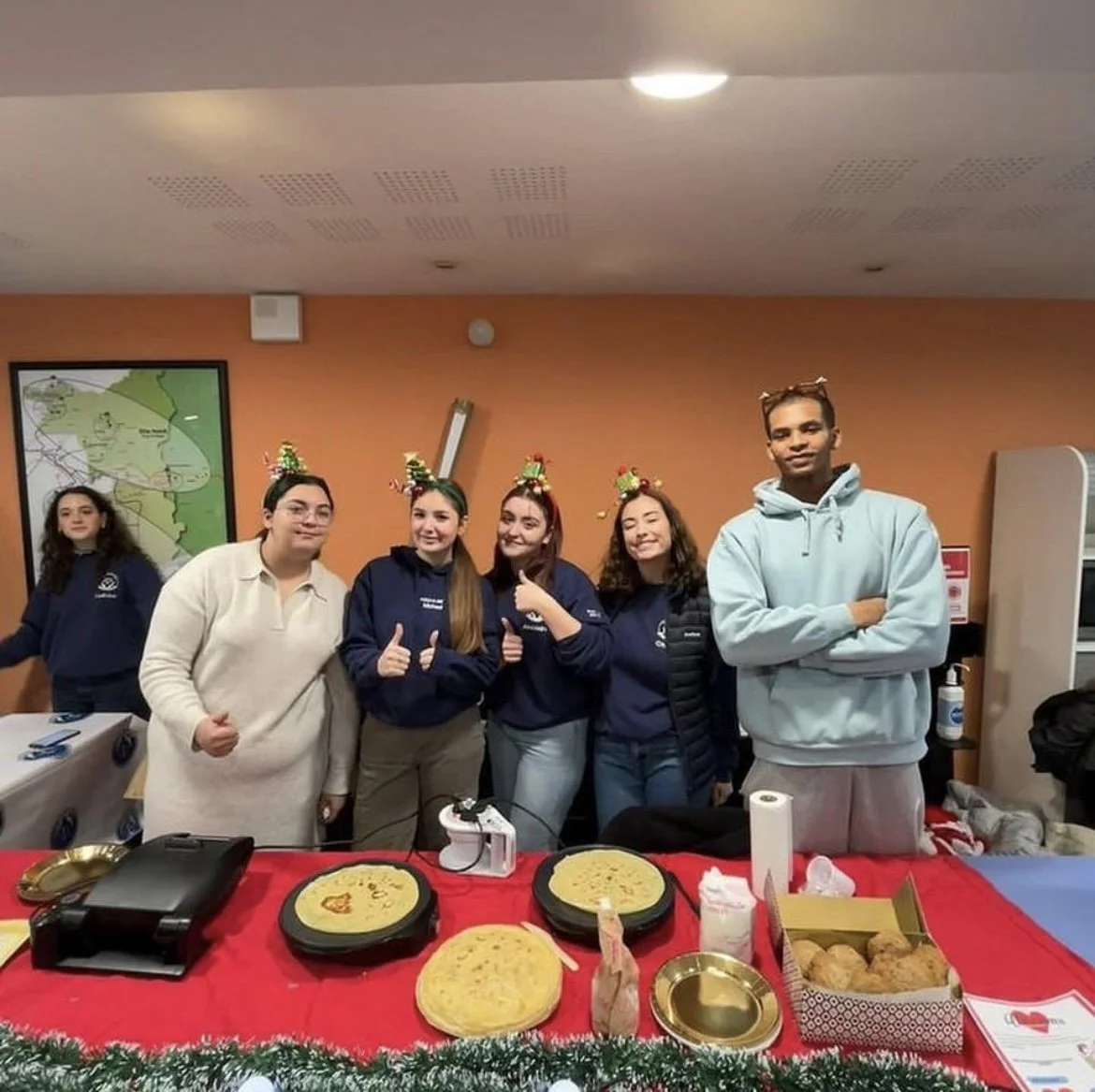 Groupe de jeunes portant des chapeaux de Noël, debout derrière une table avec des crêpes, des plats et des desserts, en intérieur, décoré pour Noël. Etudiants de Paris Assas action au profit de l'association unissons les coeurs 
