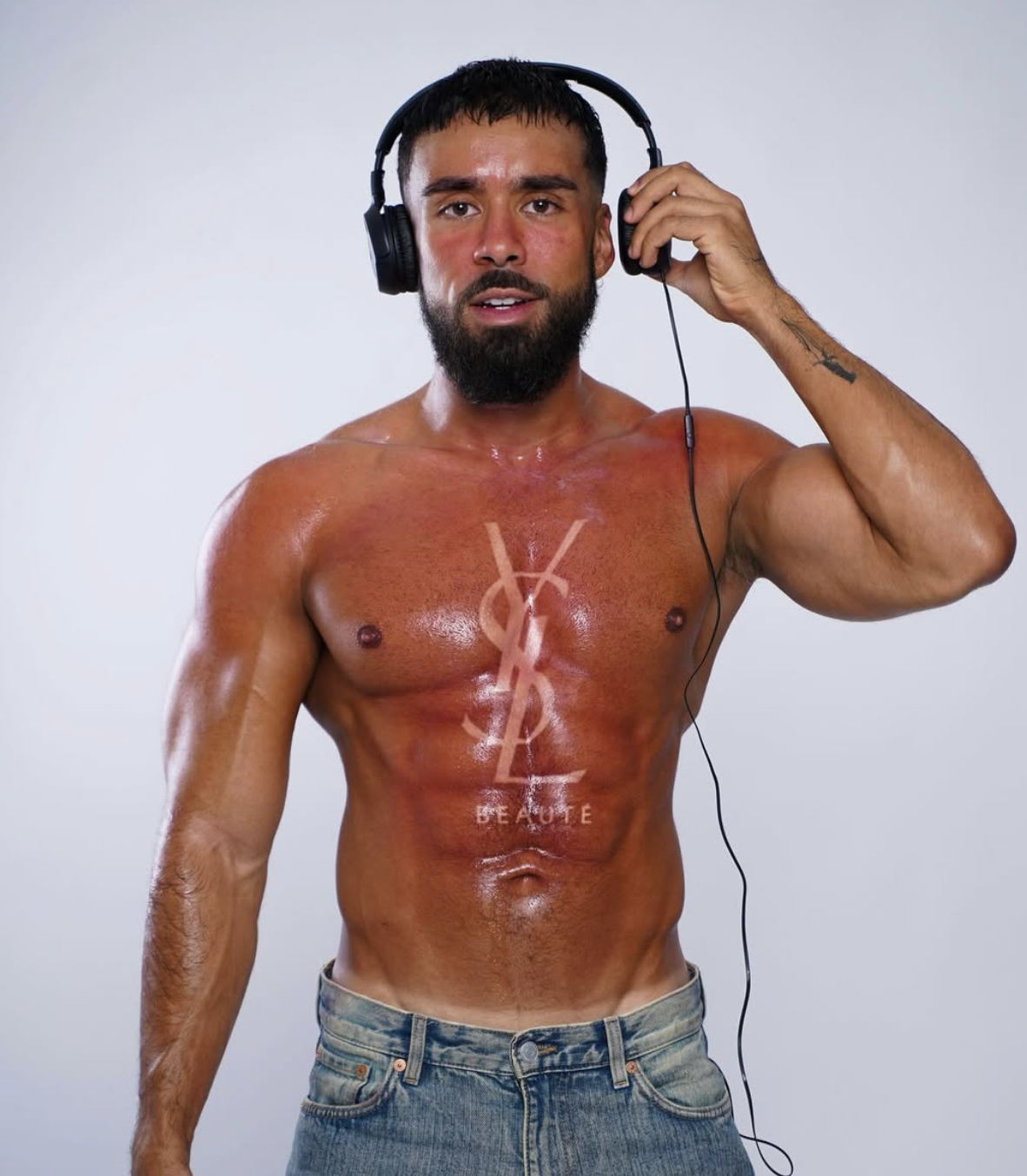 Homme chauve avec barbe, torse nu, portant des écouteurs, avec un tatouage de logo Yves Saint Laurent, et un autre tatouage sur le bras, pose devant un fond gris clair.