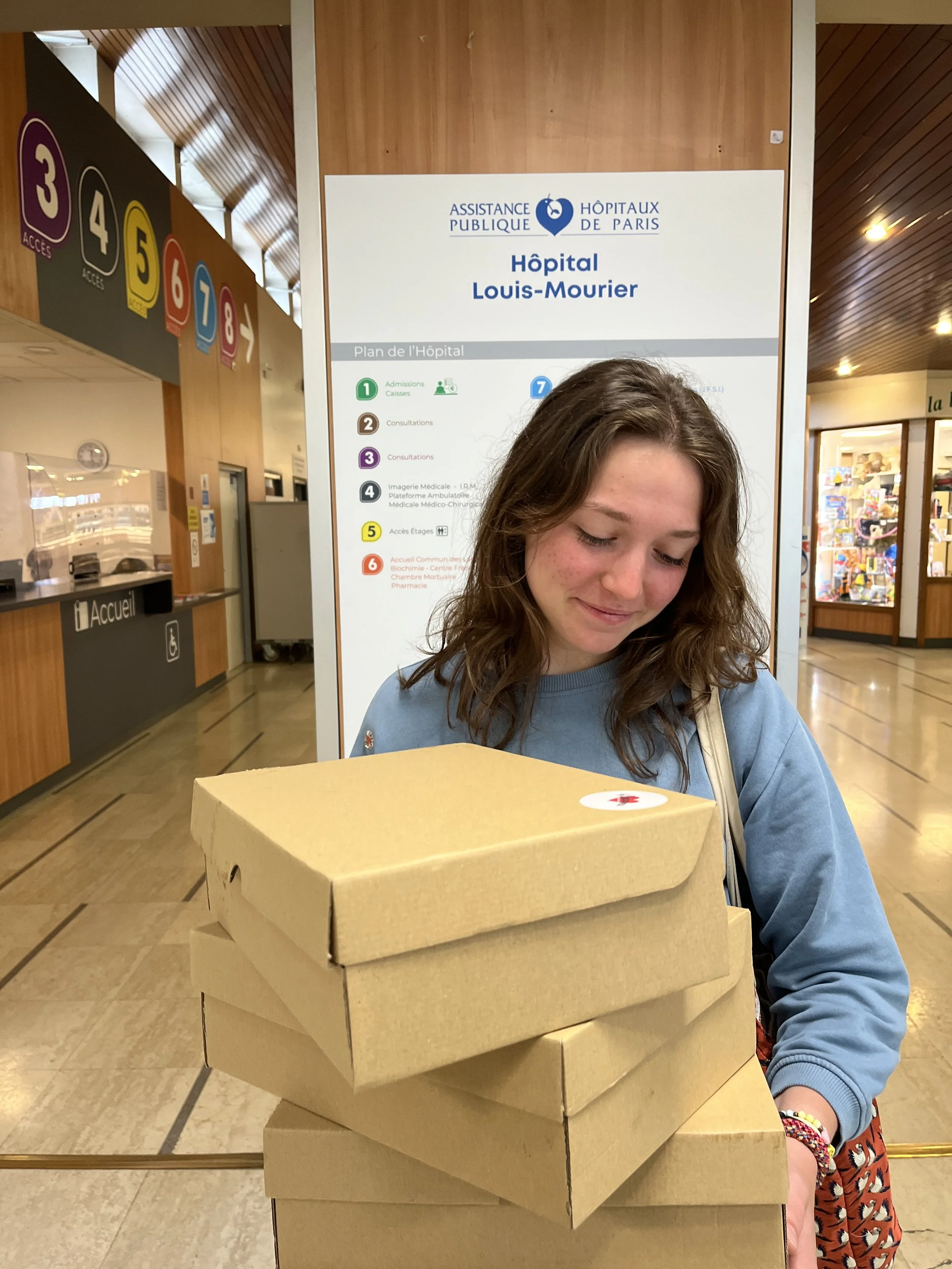 Jeune fille avec des cheveux bruns, portant un sweat bleu, posant derrière une pile de boîtes à pizza en carton dans un hall d'hôpital ou un centre médical.