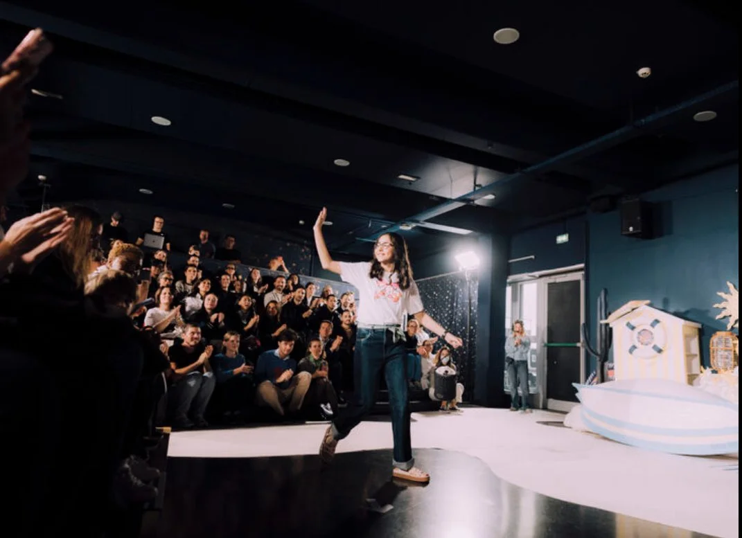 Une jeune femme marche sur scène en saluant, entourée d'un public enthousiaste dans une salle sombre avec décorations marines.