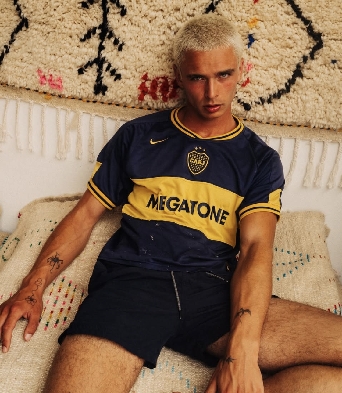 Jeune homme aux cheveux blonds courts, portant un maillot de football jaune et bleu, assis sur un canapé à côté d'un tapis suspendu.