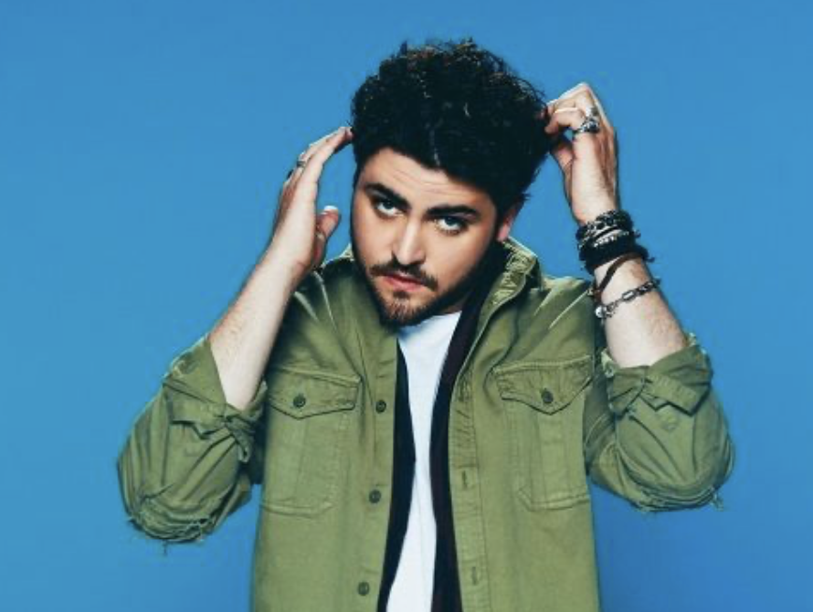 Un homme avec des cheveux bouclés, portant un manteau vert, pose contre un fond bleu.