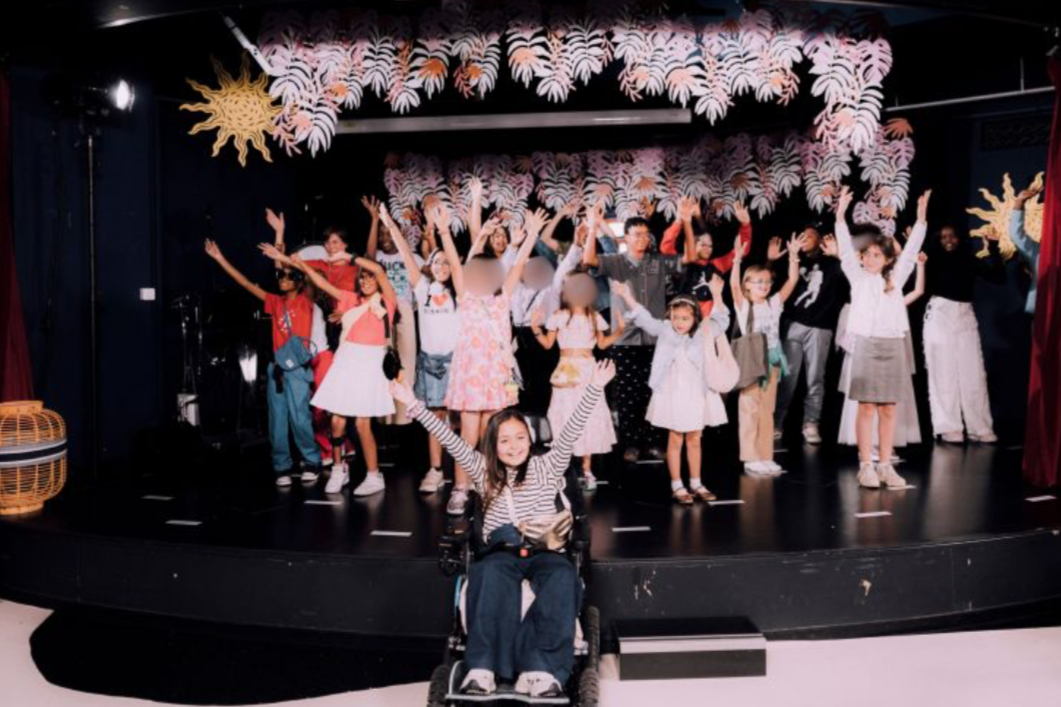 Groupe d'enfants et d'adultes sur scène, avec décor coloré de motifs floraux et de soleil en papier, certains enfants en fauteuil roulant, tous levant les bras en signe de célébration ou de joie.