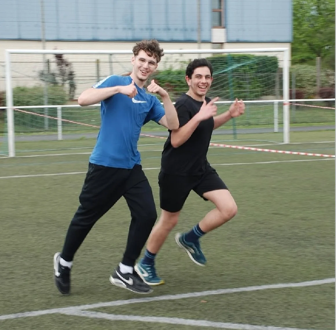 Deux jeunes hommes souriants font du jogging sur un terrain de sport en extérieur, l'un portant un t-shirt bleu et l'autre un t-shirt noir.