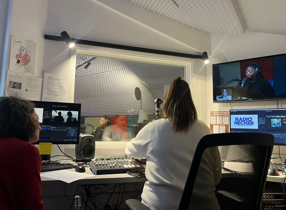 Salle d'enregistrement radio avec trois personnes, deux dans le studio et une au contrôle. Équipement audio et écrans, avec une femme parlant dans un micro.